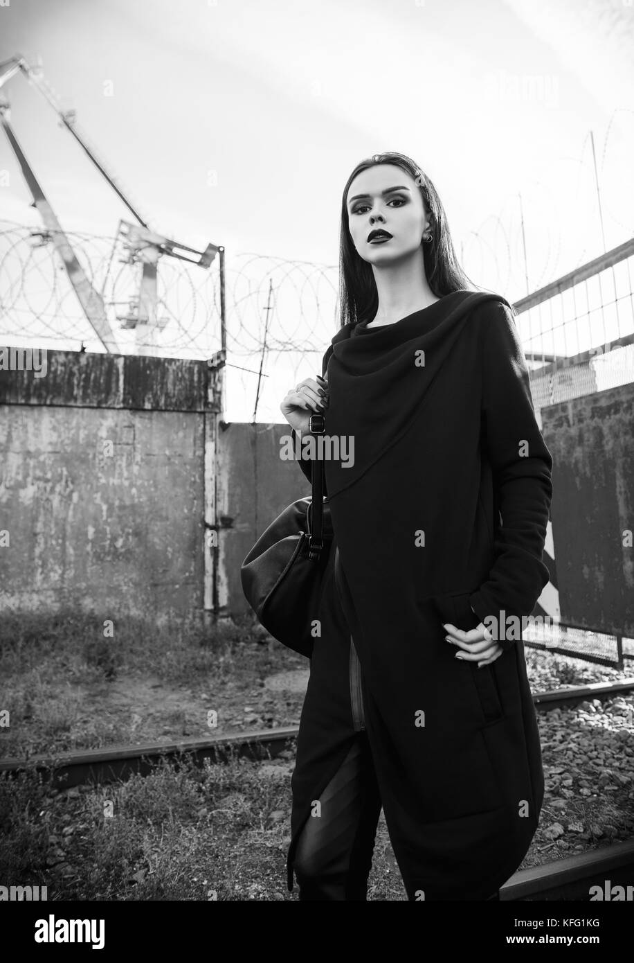Portrait of the beautiful rock girl (informal model) in tunic and leather pants standing in industrial area. Black and white Stock Photo