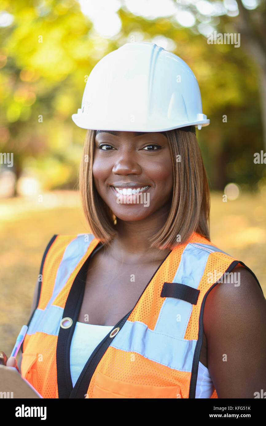 A black female construction worker wearing a white hat and orange vest working outside a city park Stock Photo