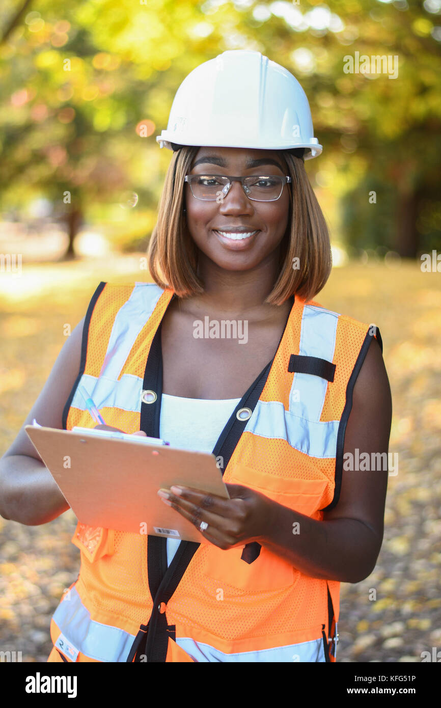 A black female construction worker wearing a white hat and orange vest working outside a city park Stock Photo
