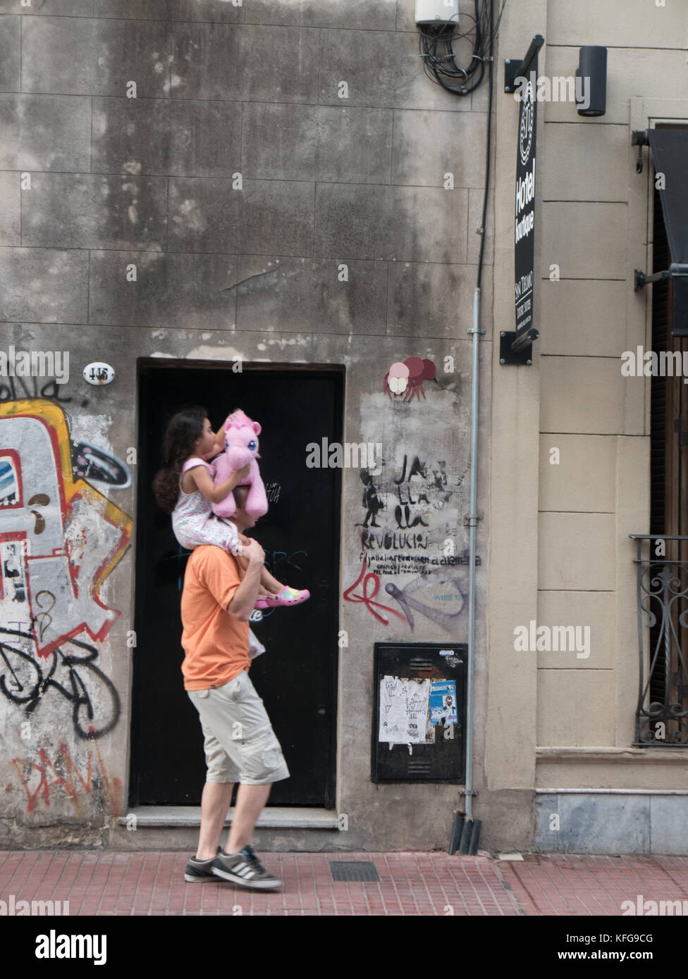 Father carrying daughter on shoulders as she holding a small pink pony stuffed animal with stone building covered with graffiti and street art. Stock Photo