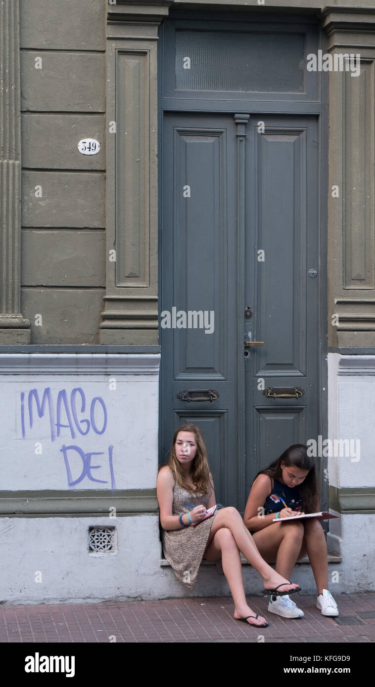 Two young women in dresses  sitting in traditional stone and wooden door entrance in San Telmo studying, writing and looking across street. Stock Photo