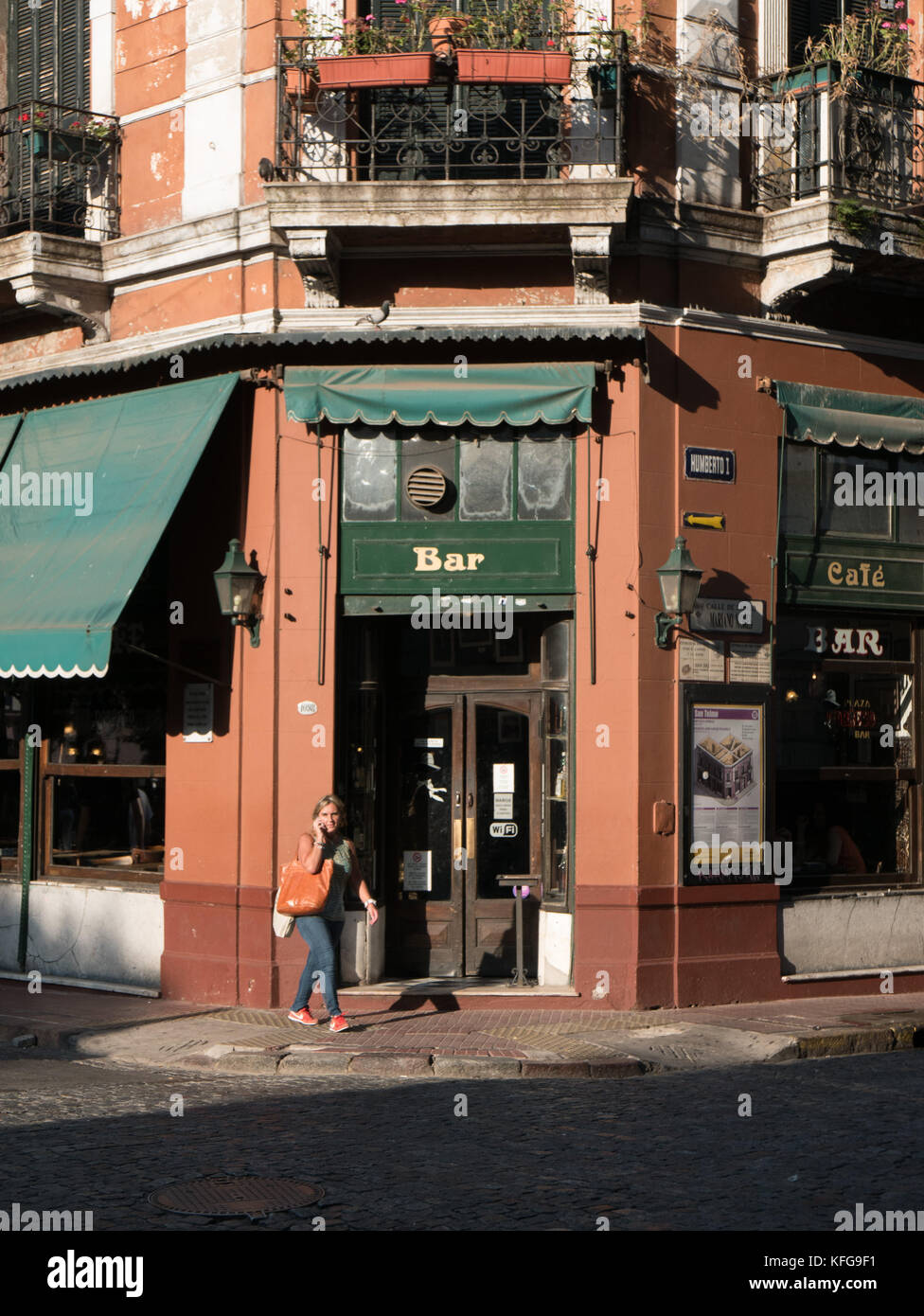 Woman walking on sidewalk in late afternoon talking on cell phone in front of old green and brown bar in San Telmo area of Bueno Aires, Argetina. Stock Photo
