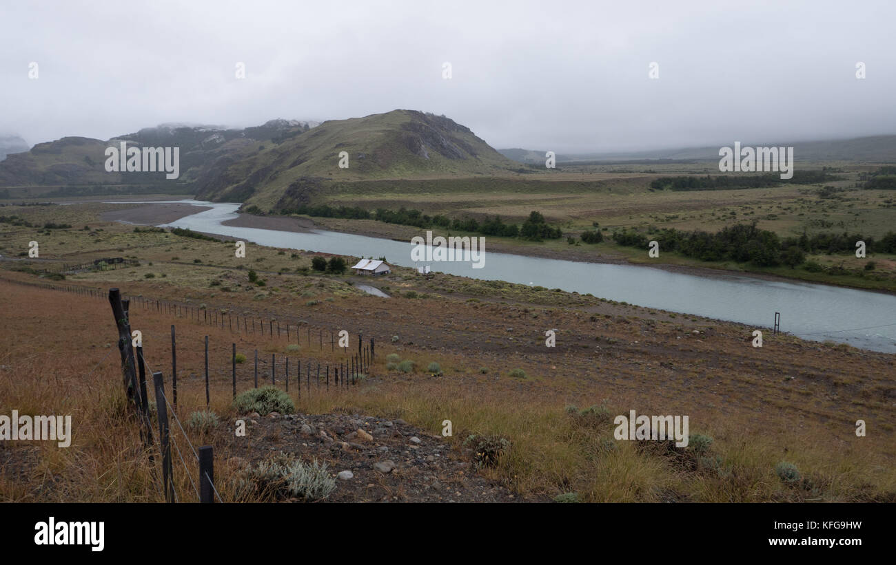 Rio de la Chinas River with misty, moody mountains in background, barb wire fence and post running down to river and green pastures in banks of river. Stock Photo