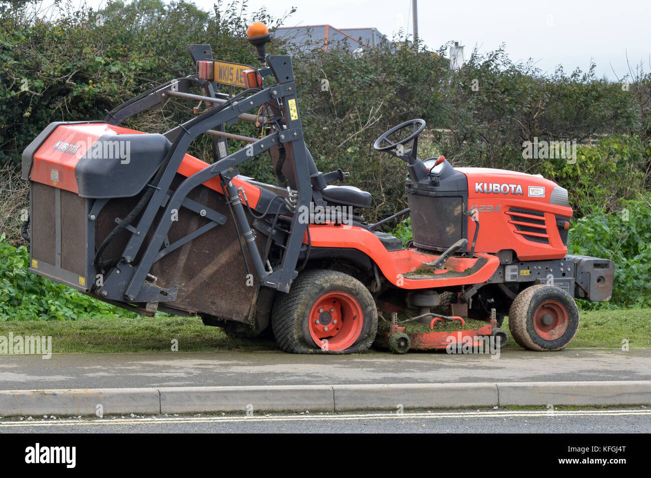 A large industrial ride-on lawnmower in orange colour parked at the side of the road on a verge with a flat tyre unable to move or mow the grass. Stock Photo