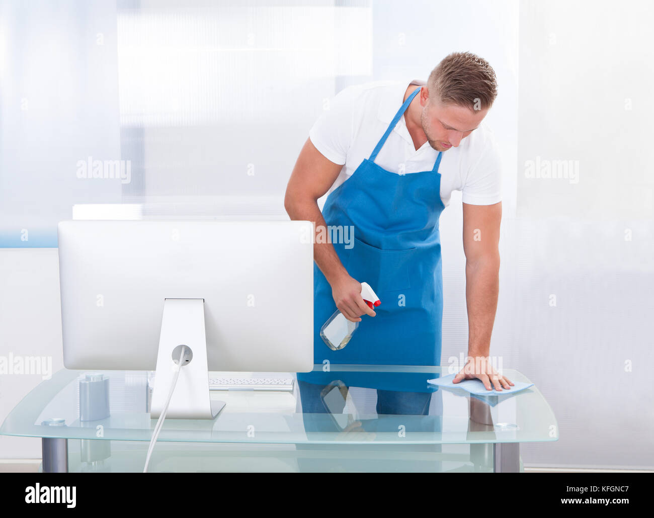 Handsome young janitor or cleaner cleaning an office spraying the top of the desk with disinfectant before the office workers start their day Stock Photo