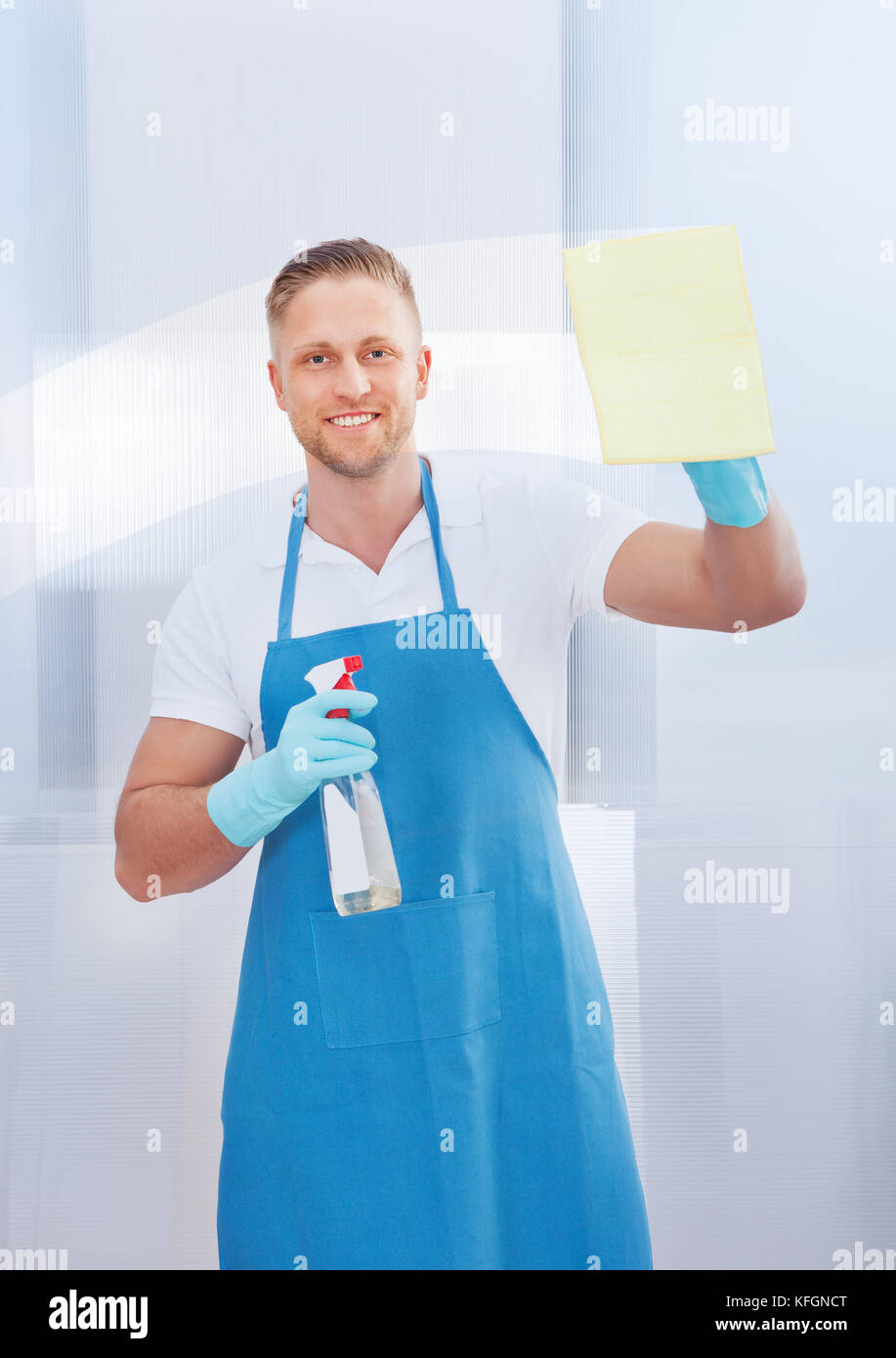 Friendly smiling male cleaner cleaning a pane of glass with a spray bottle and cloth in an office building Stock Photo