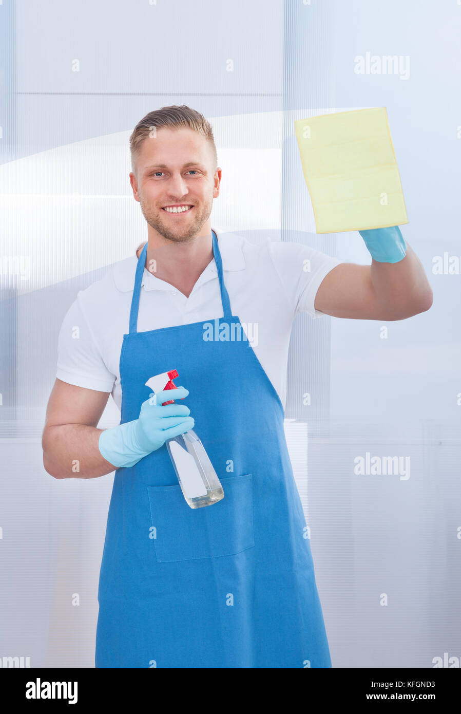 Friendly smiling male cleaner cleaning a pane of glass with a spray bottle and cloth in an office building Stock Photo