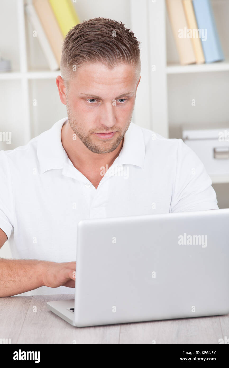 Young man in a short sleeved shirt sitting working at home on a laptop in a home office Stock Photo
