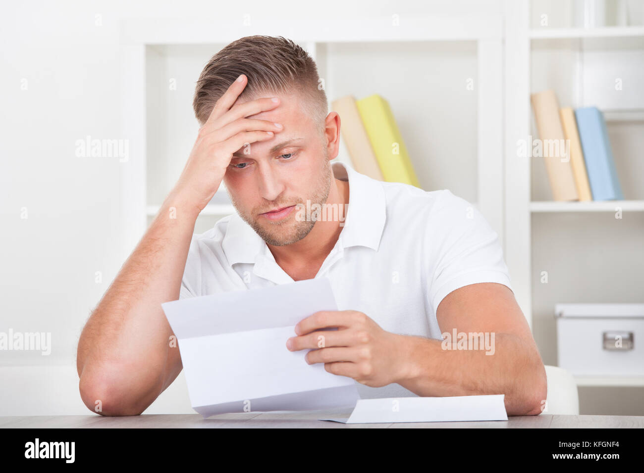 Businessman sitting in an office reacting in shock to the contents of a letter that he is reading raising his hand to his mouth Stock Photo