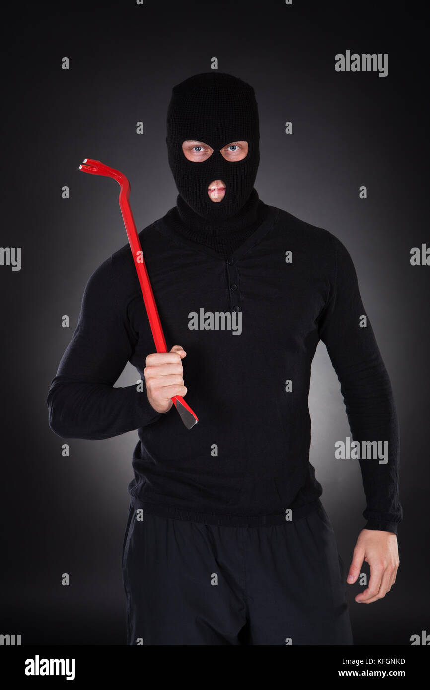 Masked thug or criminal with a crowbar raising it threateningly as he advances out of the darkness  conceptual of crime and violence Stock Photo