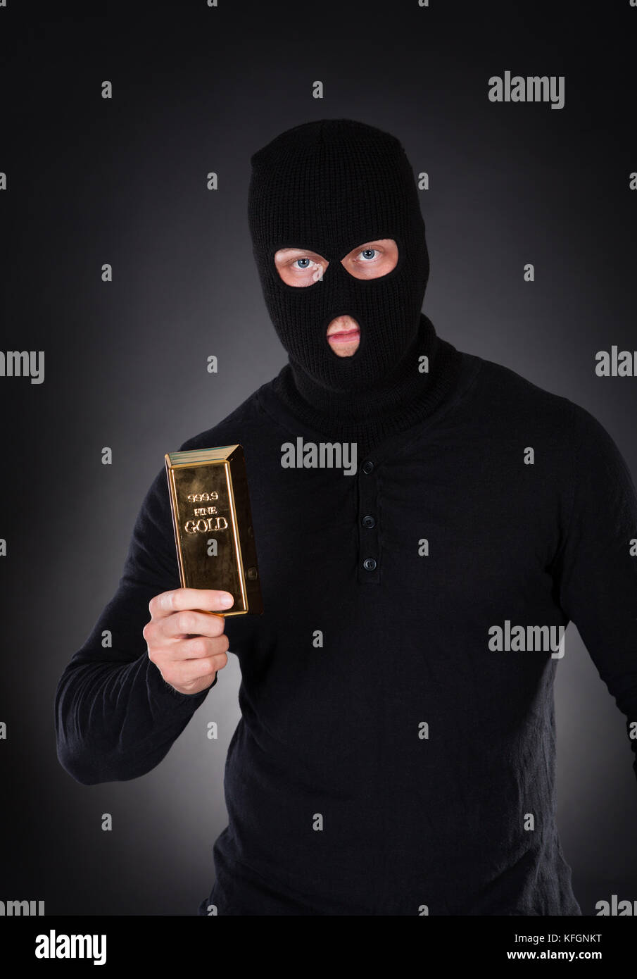 Robber disguised in a black balaclava holding a gold bullion bar as he makes his getaway from a heist with the loot through the darkness Stock Photo