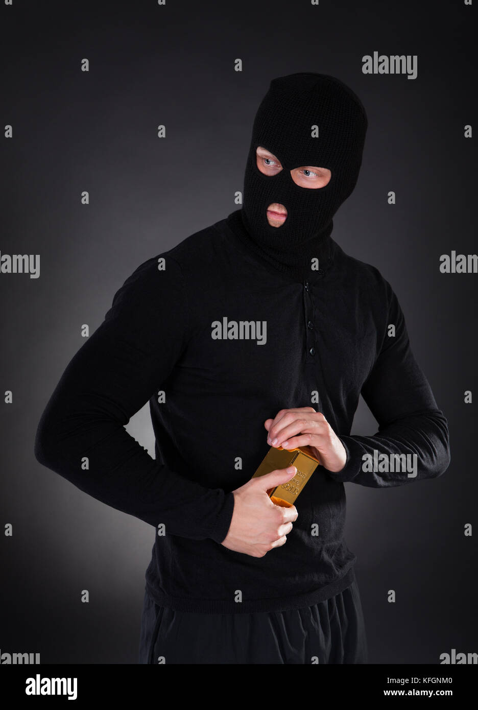 Robber disguised in a black balaclava holding a gold bullion bar as he makes his getaway from a heist with the loot through the darkness Stock Photo