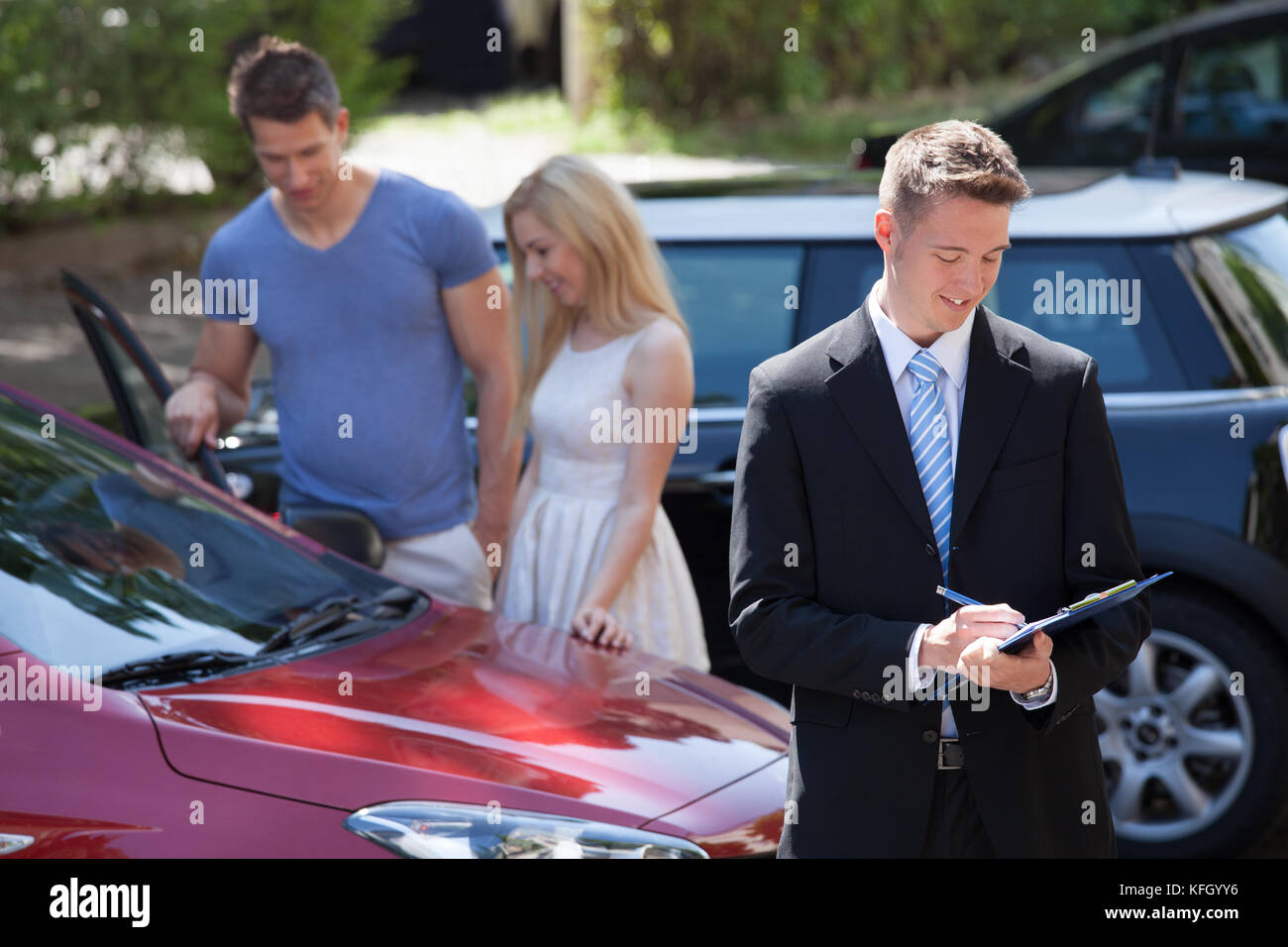 Young salesman writing on clipboard with couple looking at new car in background Stock Photo