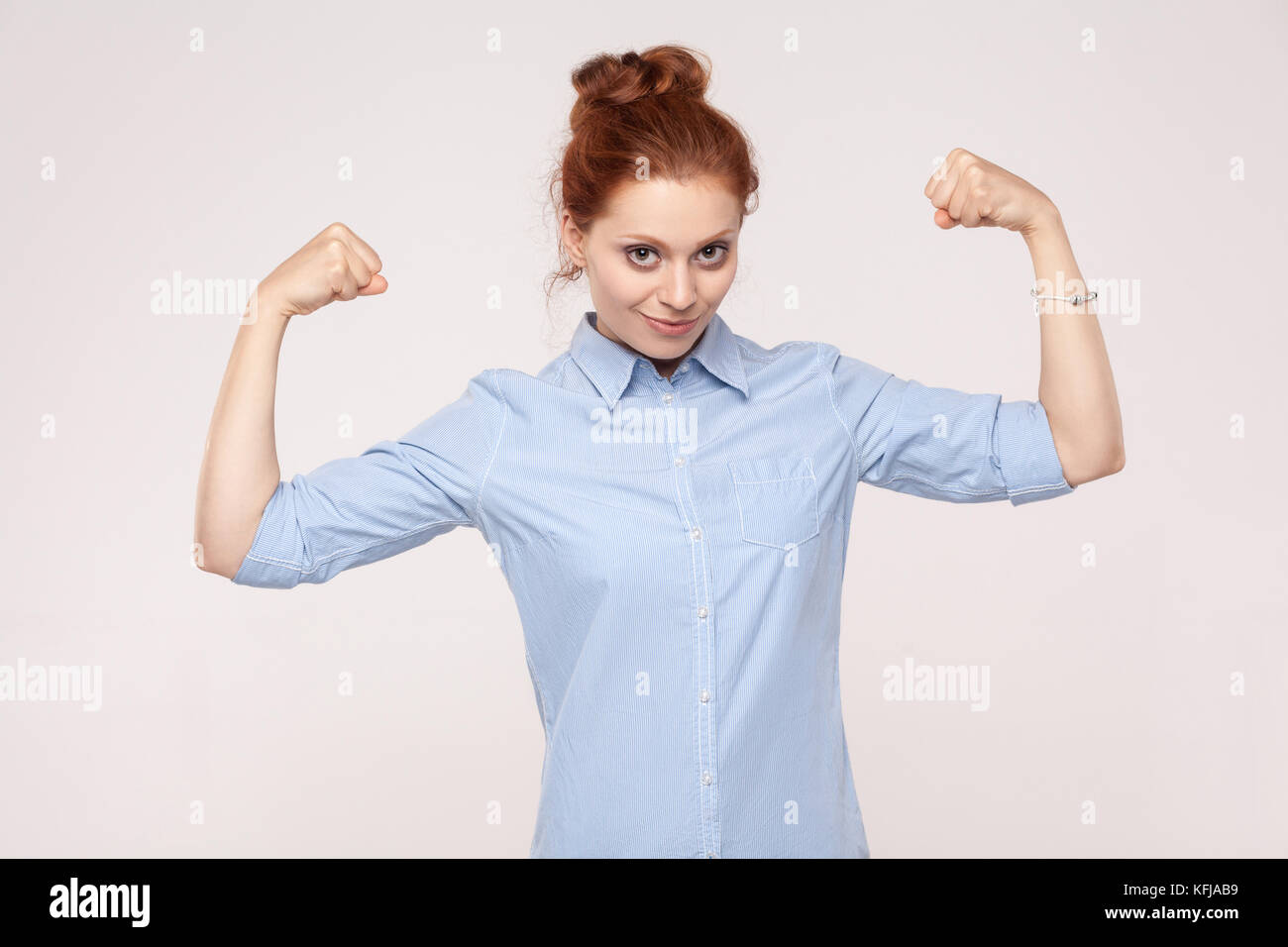 Beautifulwoman posin looks like bodybuilder, showing biceps, looking at camera and smiling. Indoor. Gray background Stock Photo
