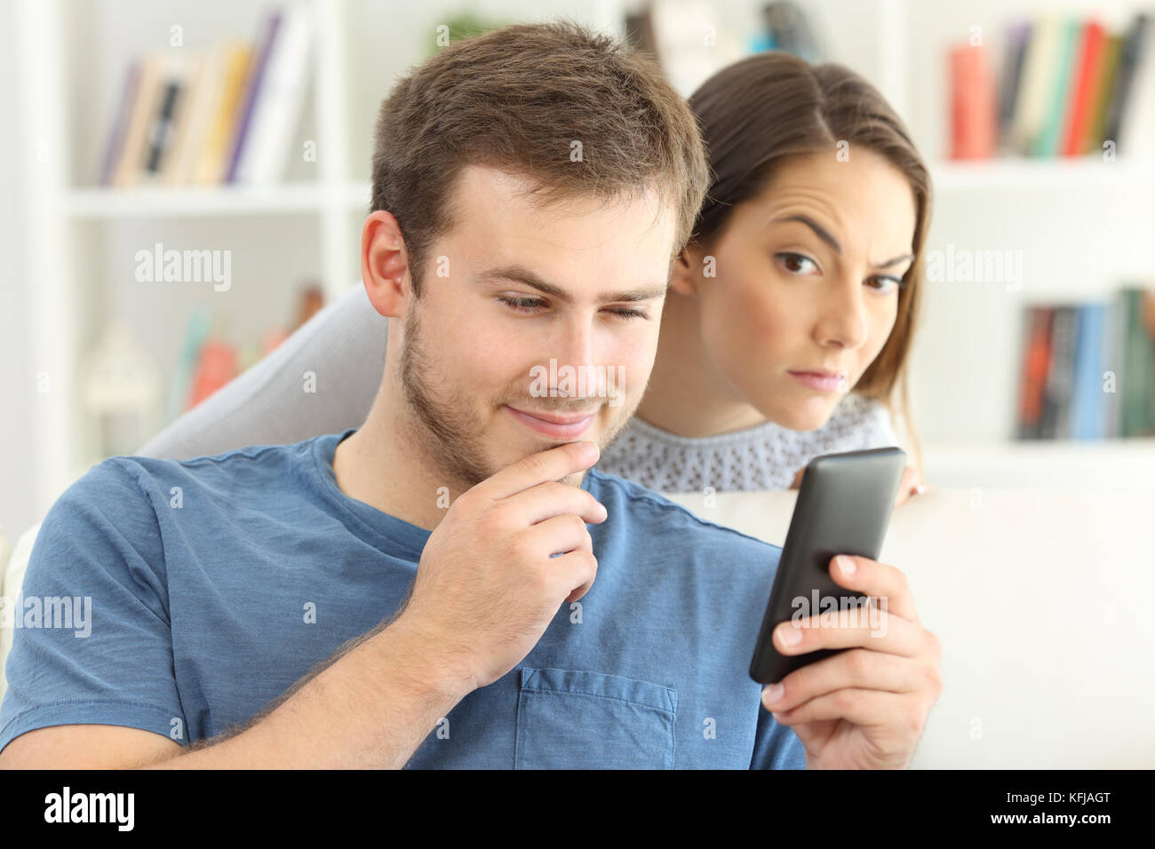 Cheater man dating on line with a smart phone and girlfriend is spying sitting on a sofa at home Stock Photo