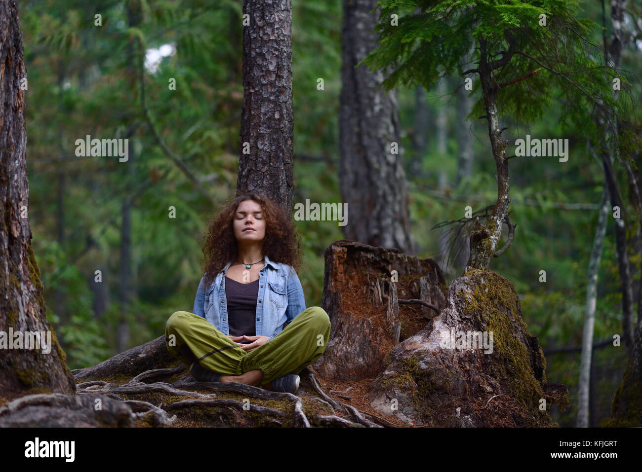 Young woman resting in a forest sitting in meditation posture leaning against a tree trunk in beautiful tranquil nature scenery Stock Photo