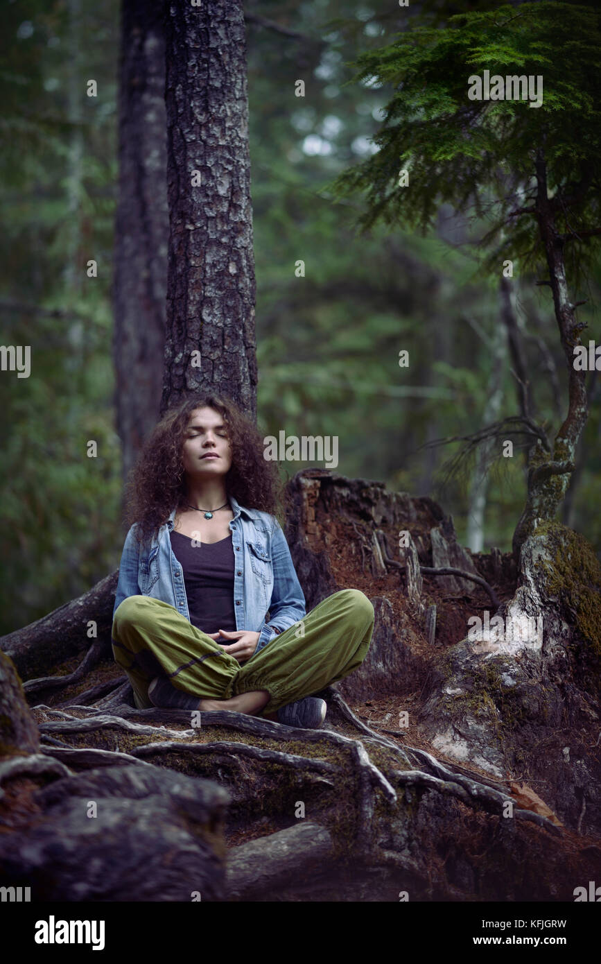 Young woman meditating in a forest sitting in meditation pose with her legs crossed leaning with her back against a tree in beautiful tranquil calm na Stock Photo