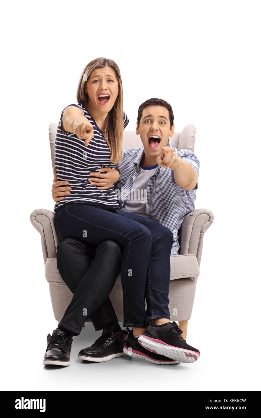 Young couple seated in an armchair pointing at the camera and laughing isolated on white background Stock Photo