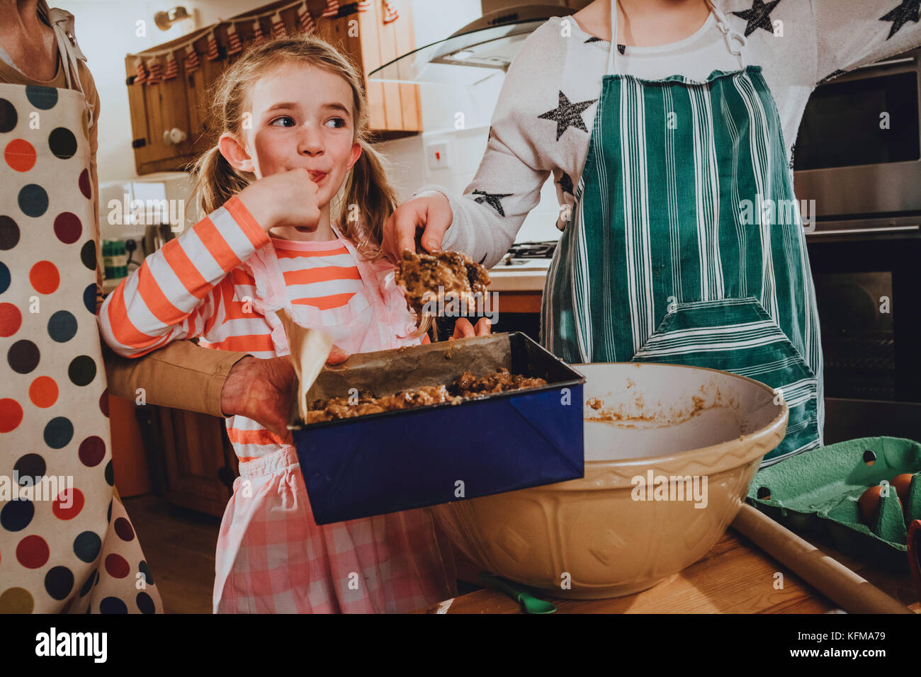 Two sisters are baking cakes with their grandmother. The youngest girl is cheekily tasting some of the cake mix on her finger. Stock Photo