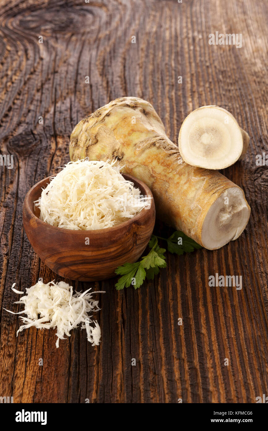Fresh and grated horseradish in wooden bowl with parsley on wooden background. Stock Photo