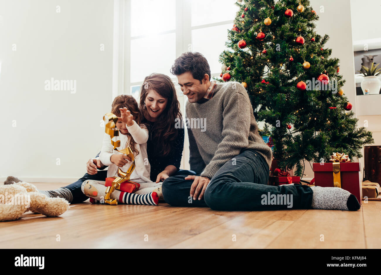 Small family having happy time together on Christmas. Young couple with child sitting beside Christmas tree opening gifts. Stock Photo