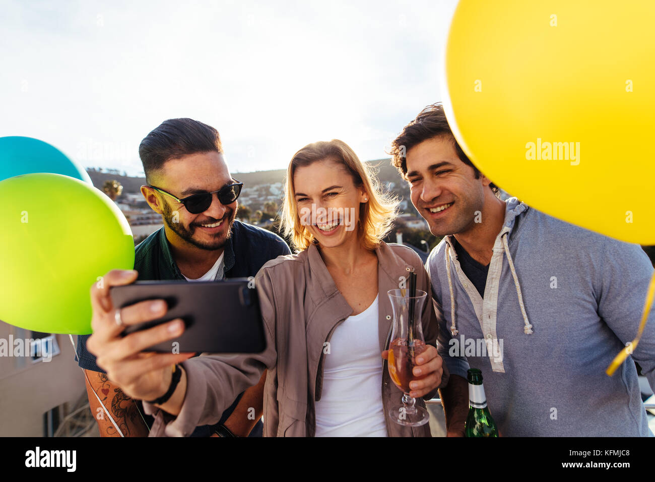 Young friends partying together taking selfie. Group of people with drinks and balloons on a rooftop party taking selfie. Stock Photo