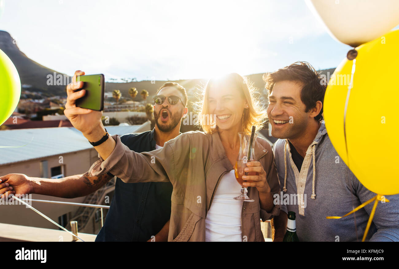 Group of friends taking selfie on a mobile phone at rooftop party. Young men and woman with drinks taking a self portrait on smart phone. Having fun o Stock Photo