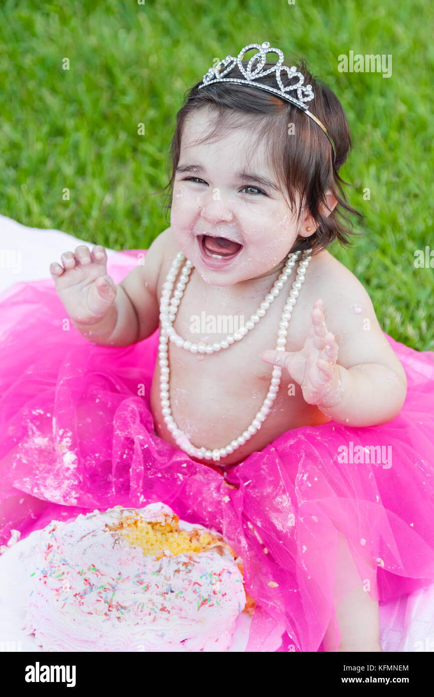Smiling happy baby toddler girl first birthday anniversary party. Ecstatic and laughing, raised hands, face dirty from pink cake. Princess costume Stock Photo