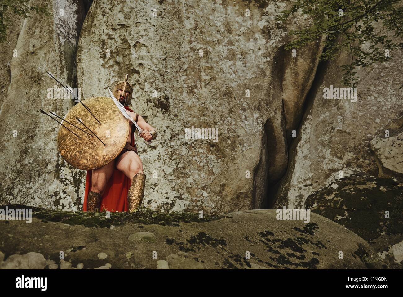 Strong warrior in gladiator armor covering with shield in arrows and posing with sword while standing on rocks. Stock Photo