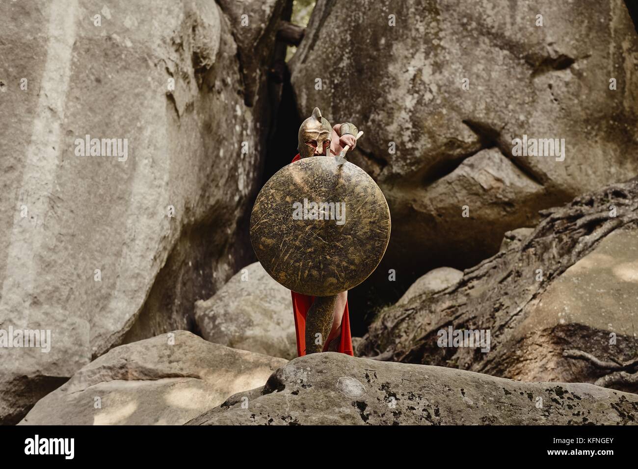 Anonymous fearless man in gladiator armor attacking with sword while covering with big metal shield on background of rocks. Stock Photo