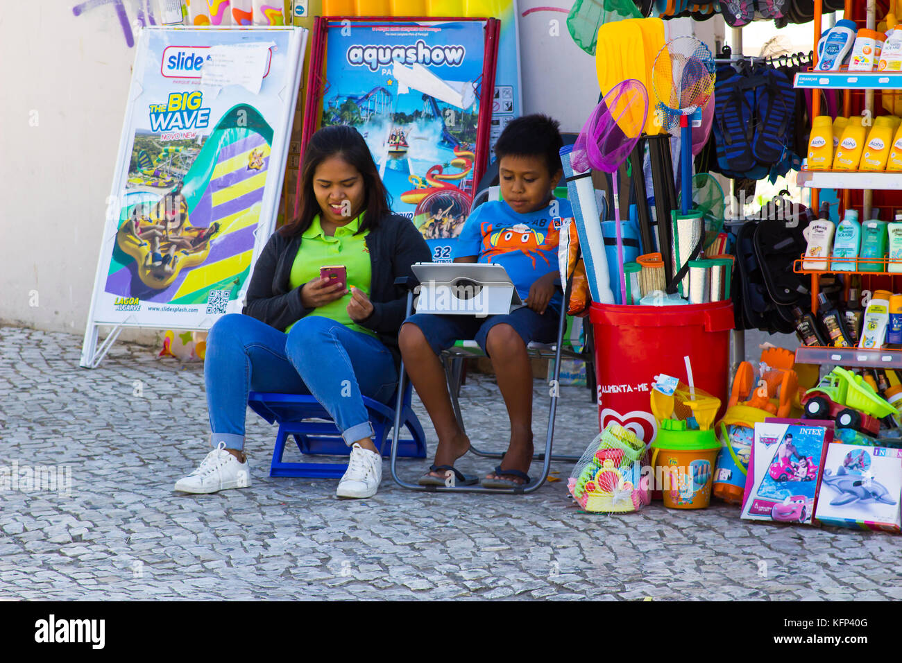 A mother and son using their mobile phone and tablet as they sit outside their small shop in Albuferia in Portuga Stock Photo