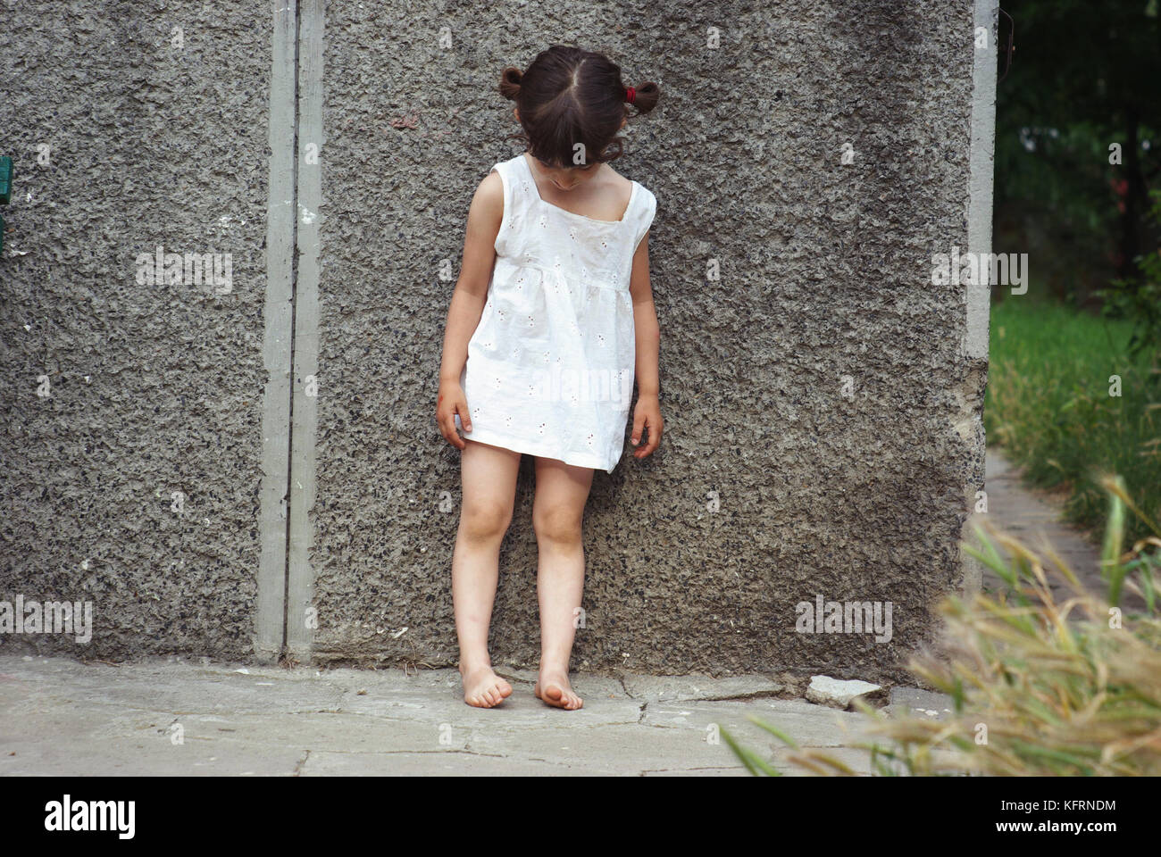 Sad little child (girl) standing against a wall, looking down. Unhappy mood, outside shot Stock Photo