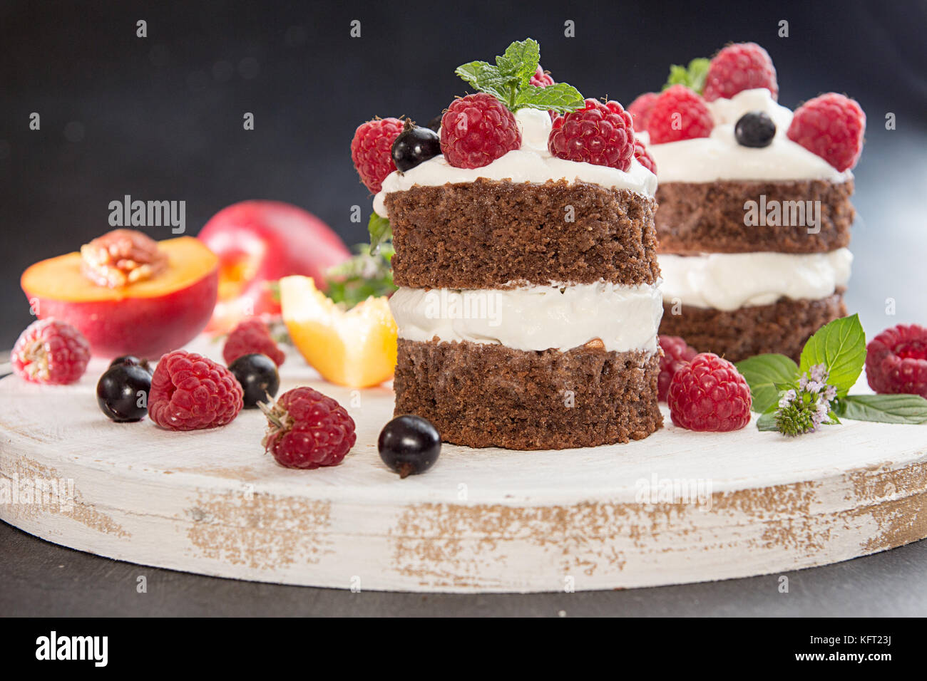 Chocolate cake decorated with raspberry, black currant, nectarine, wipped cream and mint on white wooden board on dark background Stock Photo