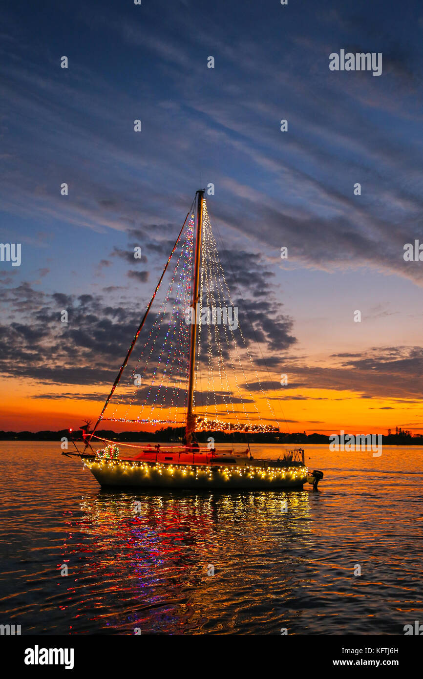 A sailboat decorated with Christmas lights passes in front of bright sunset Stock Photo