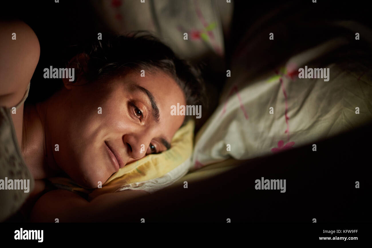 Smiling woman using tablet in bed before sleep. Bright light from digtal tablet on girl face in bed Stock Photo