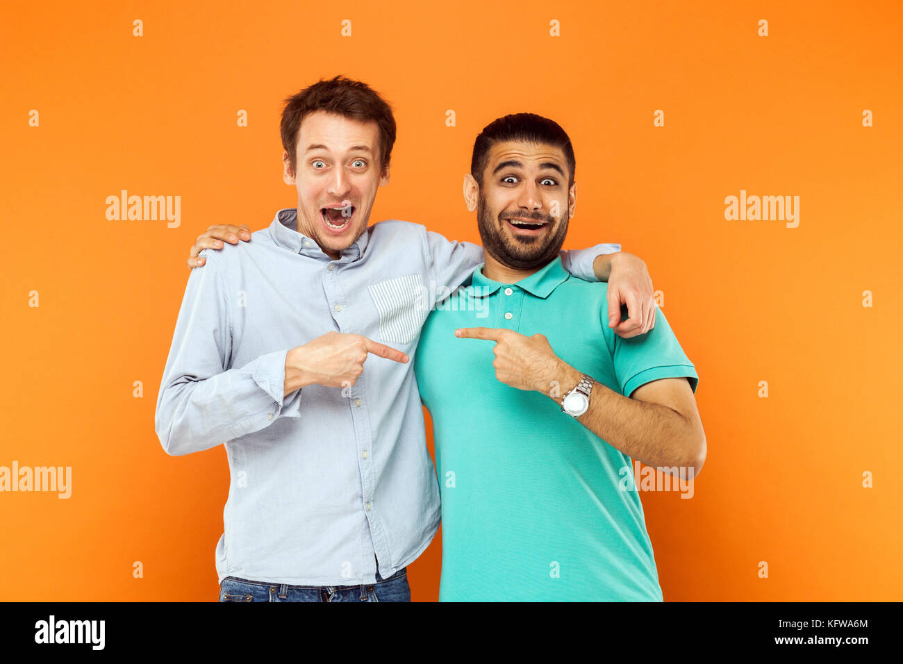 Two best friends hugging, pointing finger each other and looking at camera with shocked face. Studio shot on orange background Stock Photo