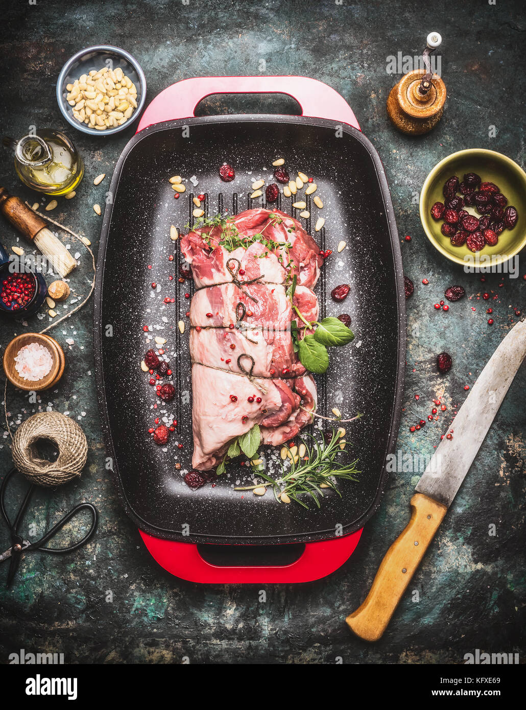 Raw Meat roast preparation in  roaster pan with cooking ingredients and seasoning on dark rustic background with butcher knife, top view Stock Photo