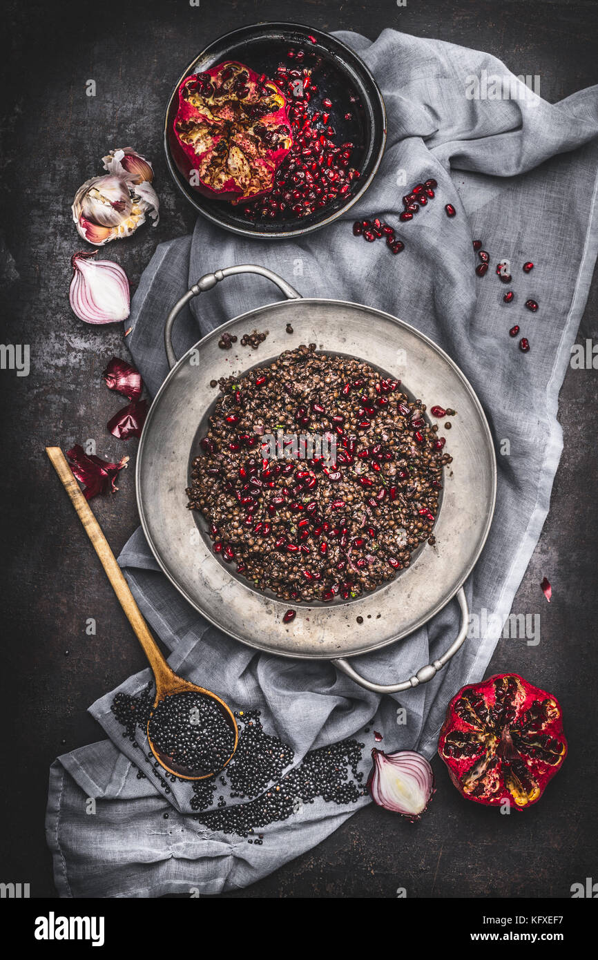 Top view of Healthy vegetarian black lentil salad with pomegranate and cooking ingredients on dark rustic background with napkin. Country dark style Stock Photo