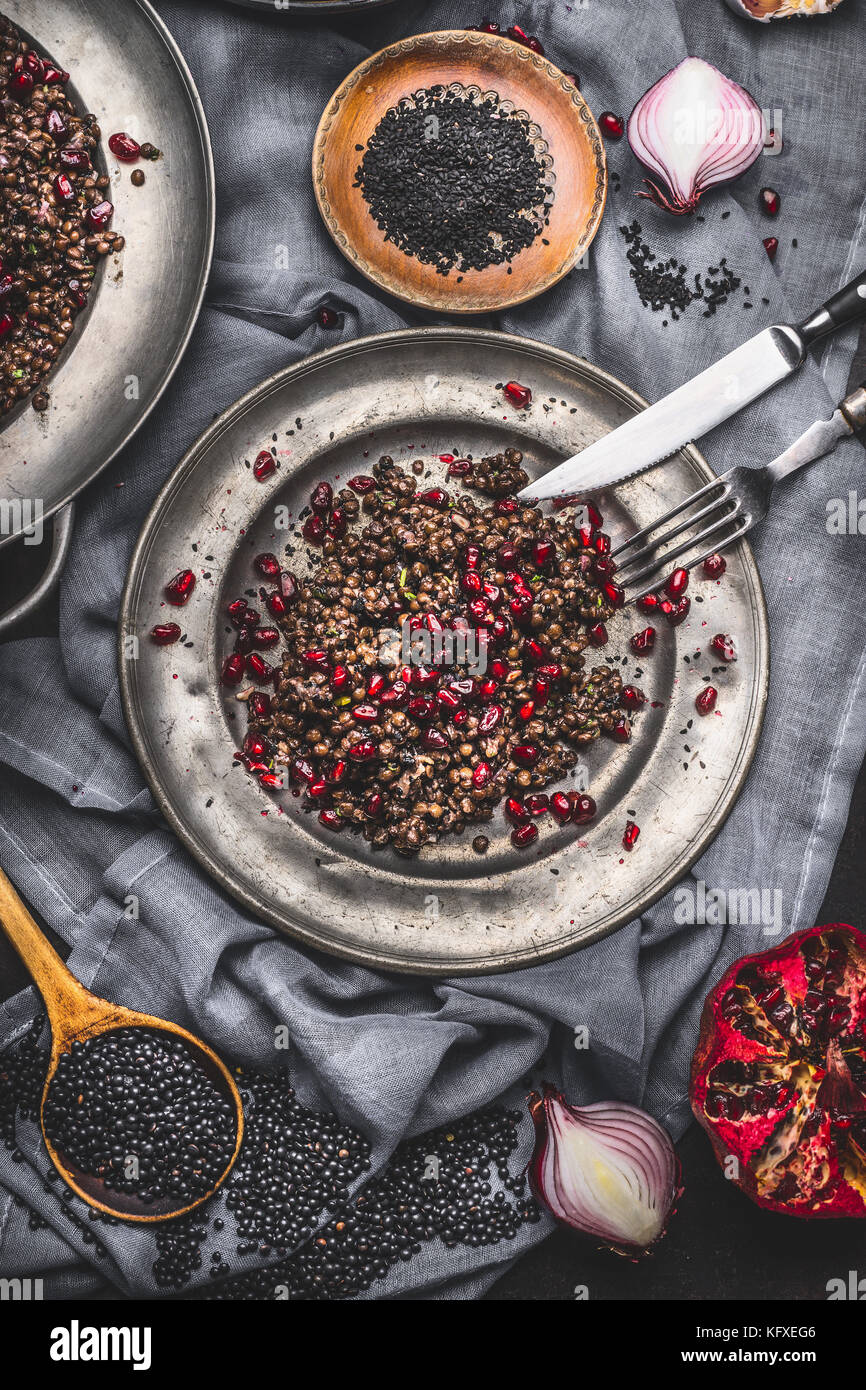 Healthy Black beluga lentil salad with pomegranate in metal plate with cutlery on dark rustic background with cooking ingredients, top view Stock Photo