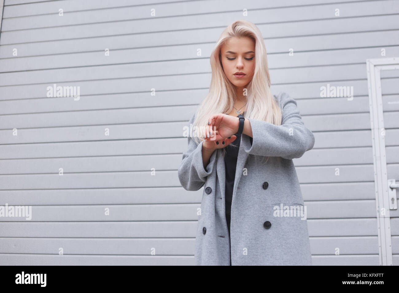 Young fashionably dressed blonde girl in a coat uses a smart bracelet standing outdoors Stock Photo