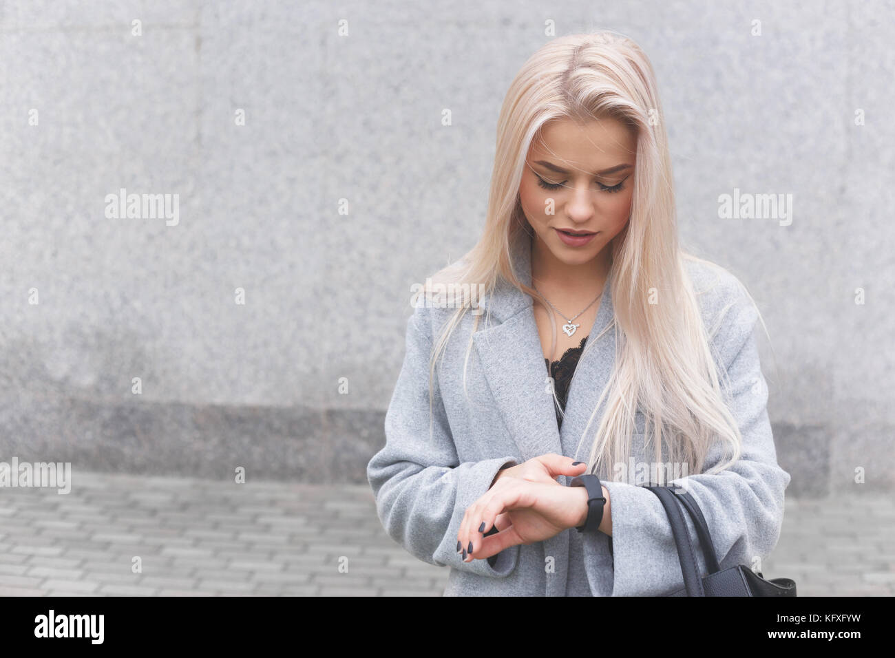 Portrait of young blondhair elegantly dressed woman in a coat uses a smart bracelet standing outdoors. Stock Photo