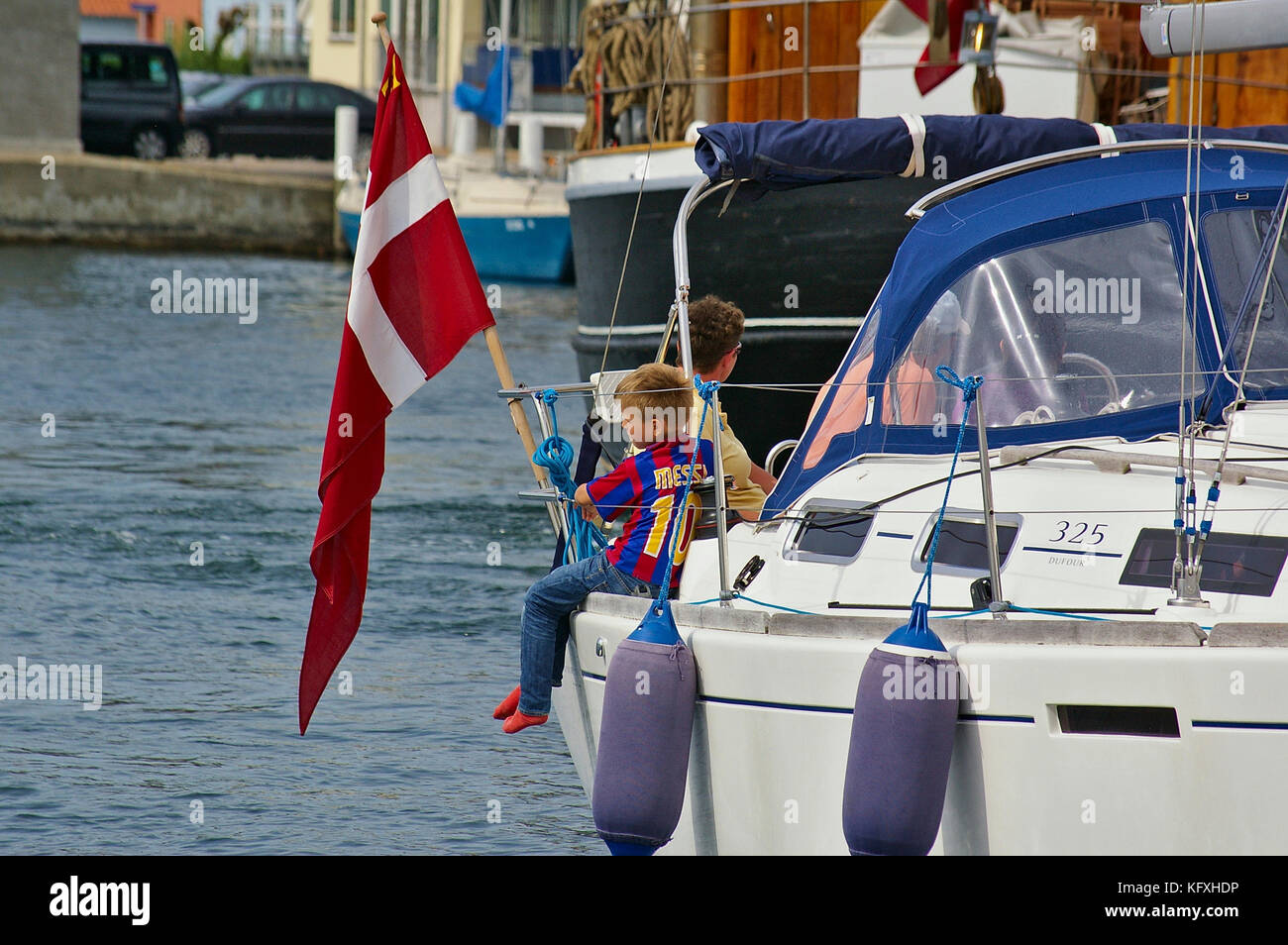 Sonderborg, Denmark - July 5th, 2012 - Young boy with soccer shirt sitting on the gunwhale of a white sailing yacht with the Danish flag next to him l Stock Photo