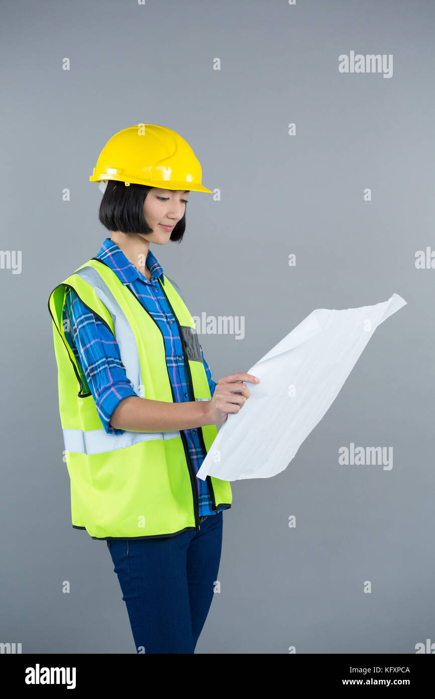 Smiling female architect looking at blueprint against grey background ...