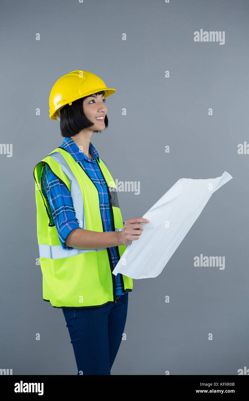 Smiling female architect looking at blueprint against grey background ...