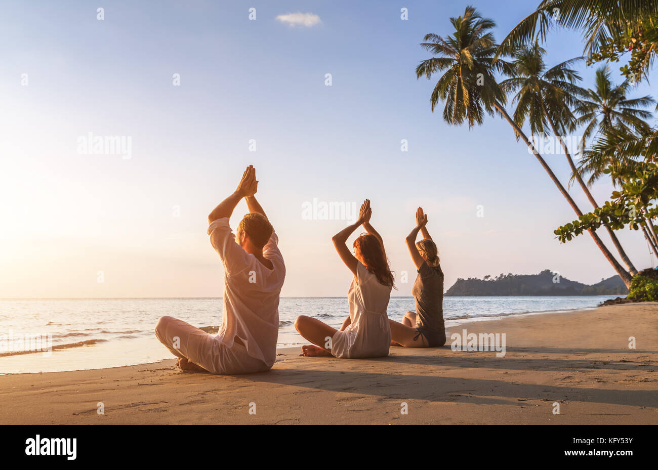 Group of three people practicing yoga lotus position on the beach for relaxation and wellbeing, warm tropical summer landscape with palm trees Stock Photo