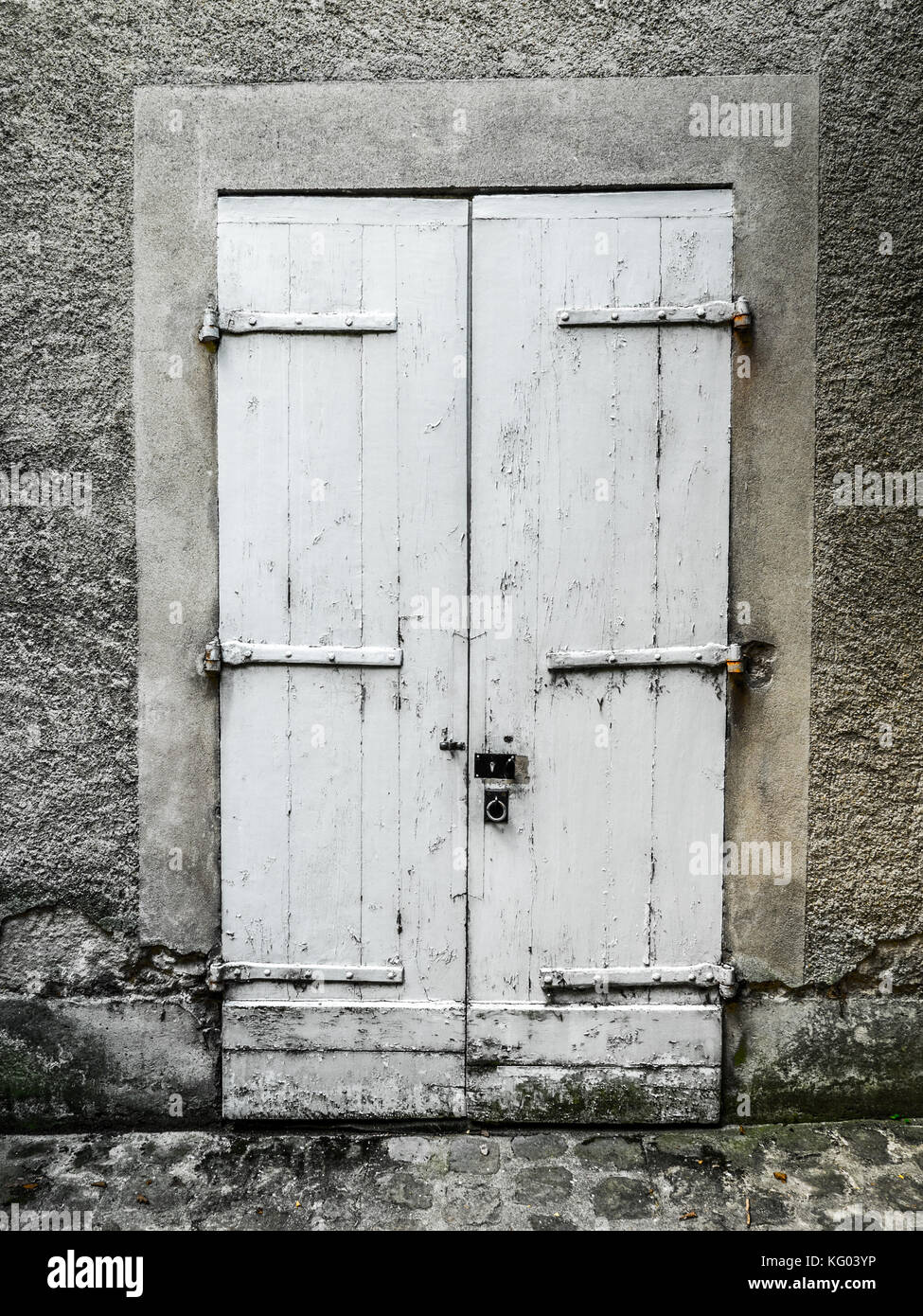 Vintage painted white wood double door with stucco frame, strap hinges, and padlock with a rustic and worn look and cobblestone road in foreground Stock Photo