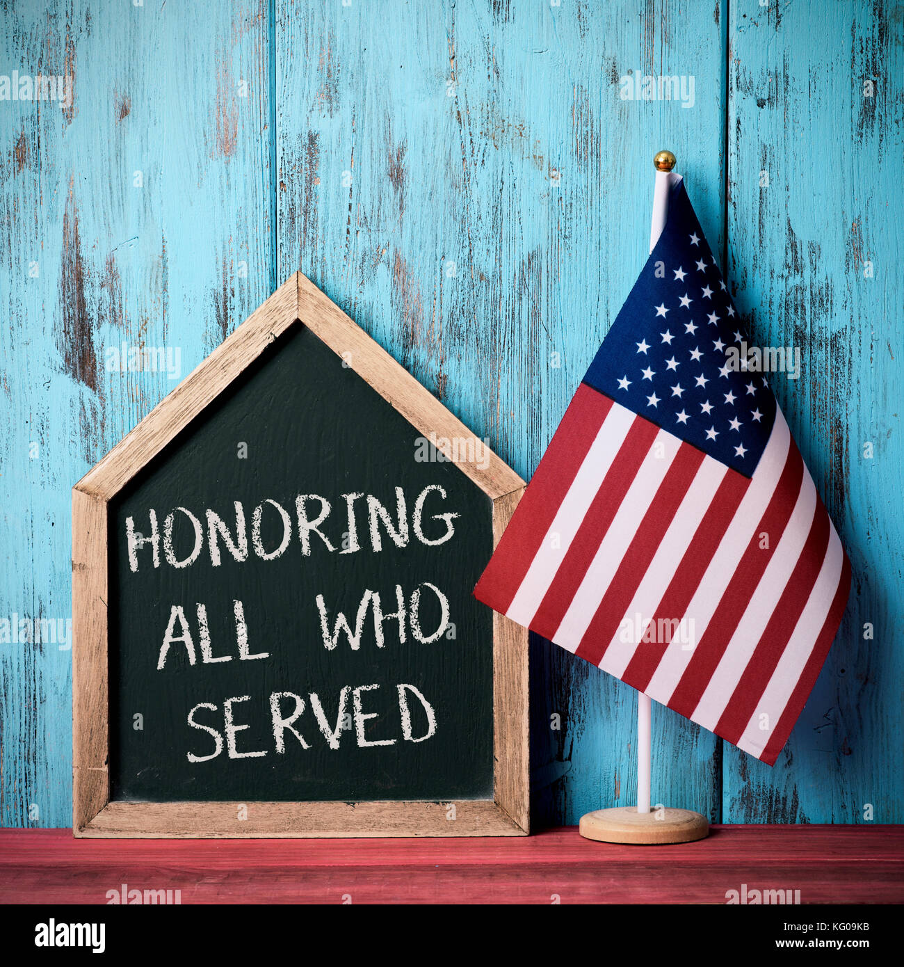 the text honoring all who served written in a house-shaped chalkboard and a flag of the United States, against a blue rustic wooden background Stock Photo