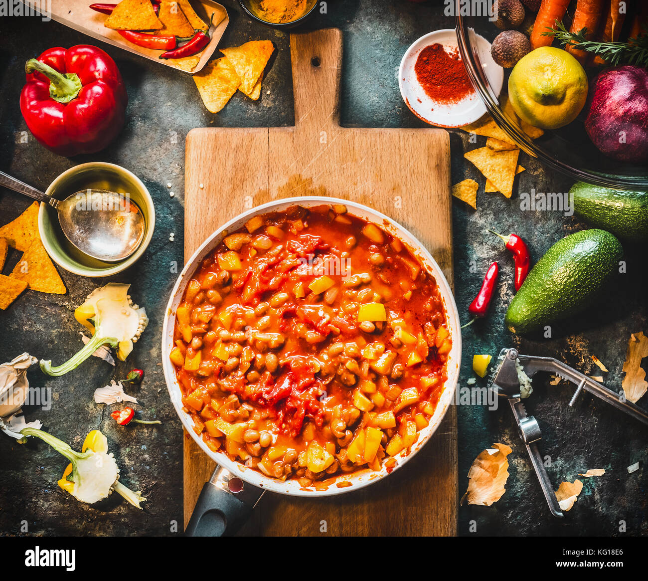 Vegetarian chili con carne dish in pan on wooden cutting board with spices and vegetables cooking ingredients on dark kitchen table background, top vi Stock Photo
