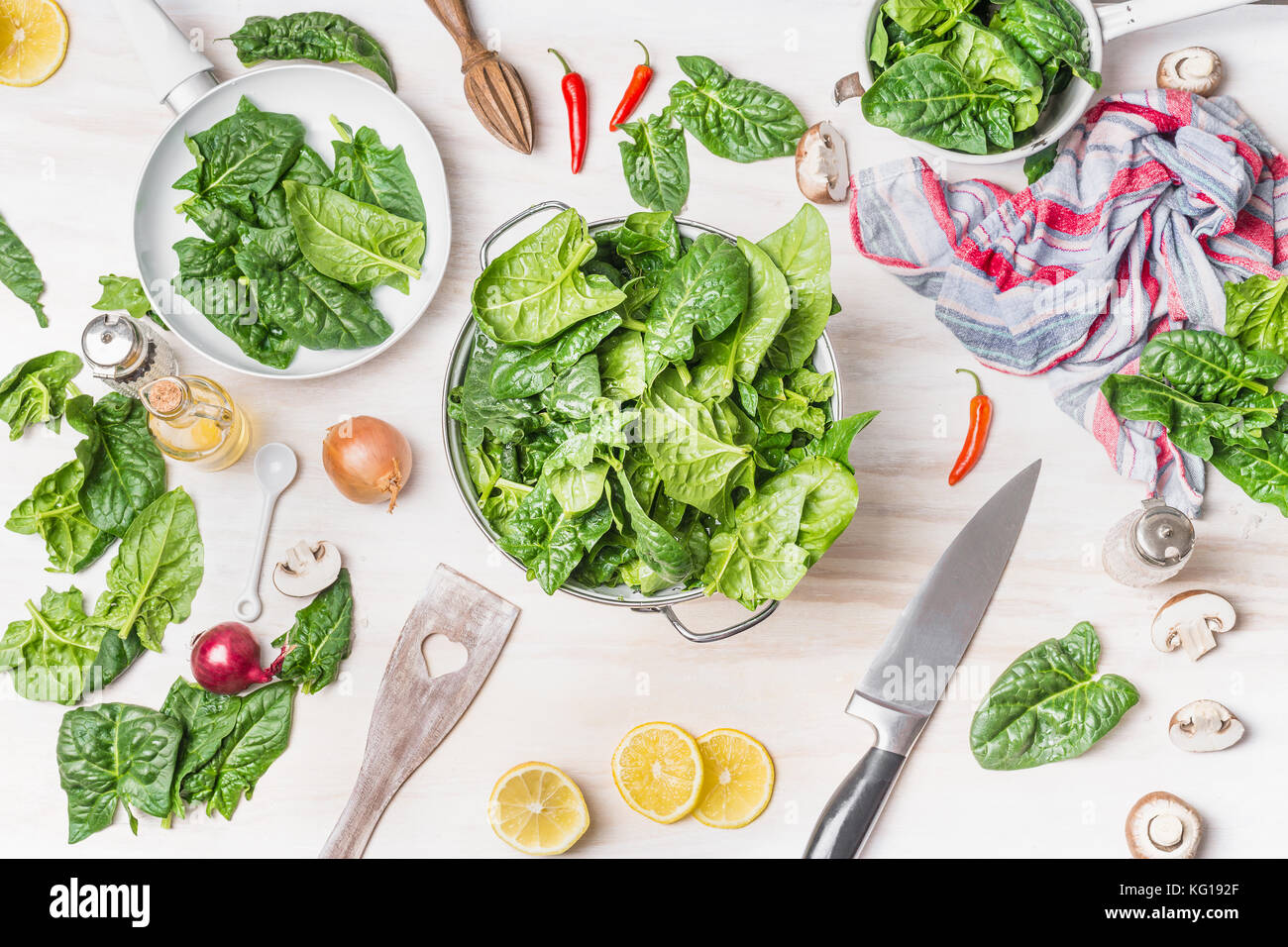 Fresh green spinach leaves in colander on white kitchen table background with cooking ingredients, spoon and knife, top view Stock Photo