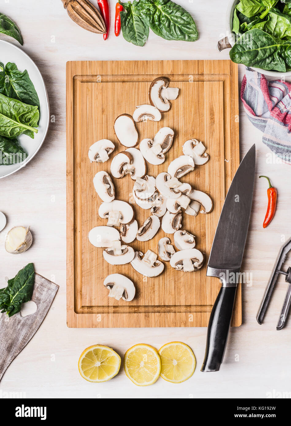 Sliced Champignons on wooden cutting board with kitchen knife and cooking ingredients: spinach and lemon on white rustic background, top view. Vegan o Stock Photo